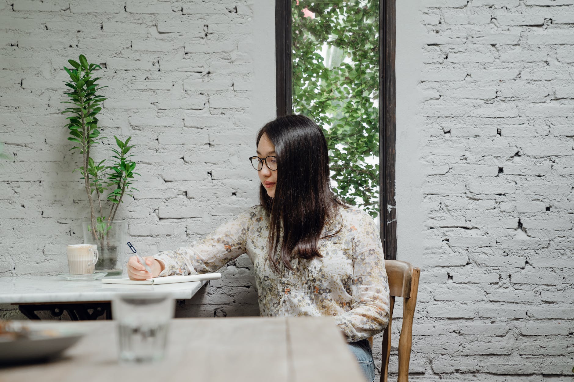 young asian woman writing in notebook sitting against white brick wall in modern cafe