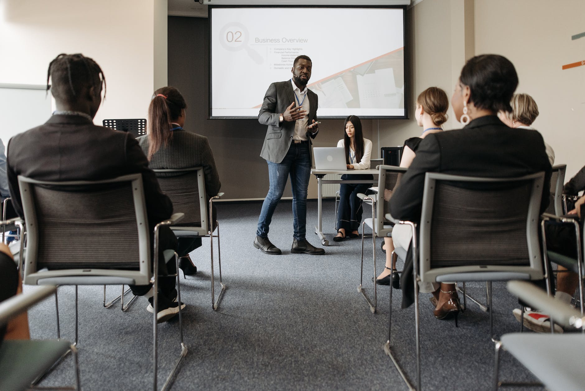 man in black suit and blue denim pants standing beside projector screen
